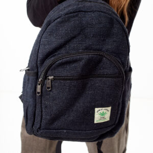 Osoba na białym tle w brązowych spodniach we wzór gheri i czarnym topie trzyma w rękach czarny plecak z konopi z wieloma zapięciami