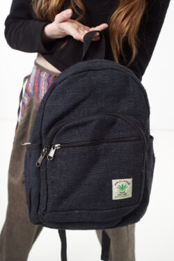 Osoba na białym tle w brązowych spodniach we wzór gheri i czarnym topie trzyma w rękach czarny plecak z konopi z wieloma zapięciami