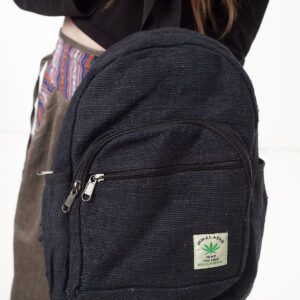 Osoba na białym tle w brązowych spodniach we wzór gheri i czarnym topie trzyma w rękach czarny plecak z konopi z wieloma zapięciami