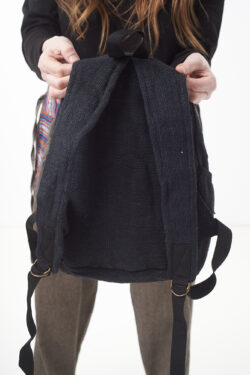 Osoba na białym tle w brązowych spodniach we wzór gheri i czarnym topie trzyma w rękach czarny plecak z konopi, pokazując jego tył