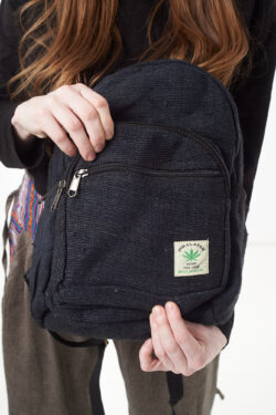 Osoba na białym tle w brązowych spodniach we wzór gheri i czarnym topie trzyma w rękach czarny plecak z konopi z wieloma zapięciami