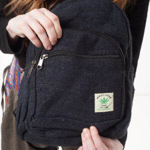 Osoba na białym tle w brązowych spodniach we wzór gheri i czarnym topie trzyma w rękach czarny plecak z konopi z wieloma zapięciami