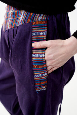 Osoba w czarnej koszulce wkłada rękę do kieszeni fioletowych spodni z wstawkami gheri na białym tle