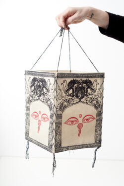 Ręka trzyma beżowy lampion z czerpanego papieru z ilustracją oczu buddy i czarnych smoków, na białym tle