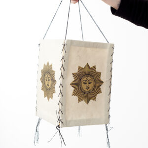 Ręka trzyma beżowy lampion z czerpanego papieru z ilustracją złotego słońca, na białym tle