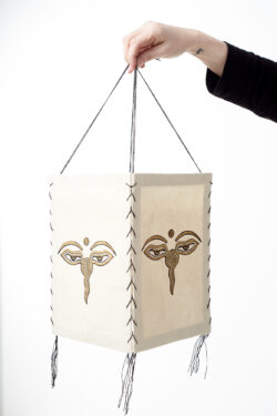 Ręka trzyma beżowy lampion z czerpanego papieru z ilustracją złotych oczu buddy, na białym tle