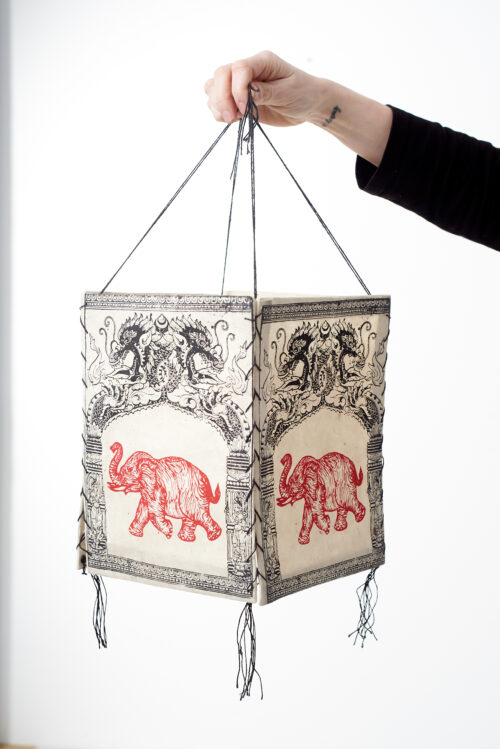 Ręka trzyma beżowy lampion z czerpanego papieru z ilustracją czerwonego słonia i czarnych etnicznych ornamentów, na białym tle