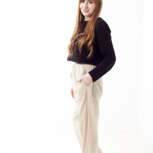 Kobieta w czarnej koszulce wkłada rękę do kieszeni luźnych beżowych spodni na białym tle