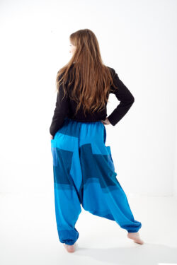 Osoba stoi tyłem na białym tle w luźnych niebieskich patchworkowych szarawarach z kieszeniami