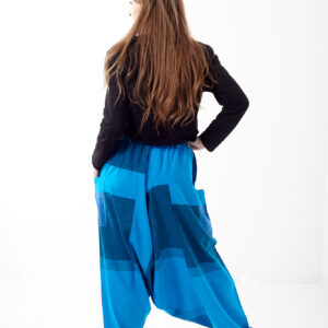 Osoba stoi tyłem na białym tle w luźnych niebieskich patchworkowych szarawarach z kieszeniami