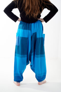 Osoba stoi tyłem na białym tle w oversize’owych niebieskich patchworkowych szarawarach z kieszeniami