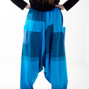 Osoba stoi tyłem na białym tle w oversize’owych niebieskich patchworkowych szarawarach z kieszeniami