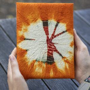 Osoba trzyma notes z pomarańczowo-bordowym wzorem tie-dye na drewnianym tle