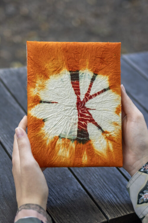 Osoba trzyma notes z pomarańczowo-bordowym wzorem tie-dye na drewnianym tle