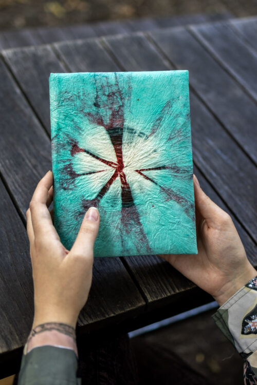 Osoba trzyma notes z turkusowo-bordowym wzorem tie-dye na drewnianym tle