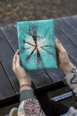 Osoba trzyma notes z turkusowo-bordowym wzorem tie-dye na drewnianym tle