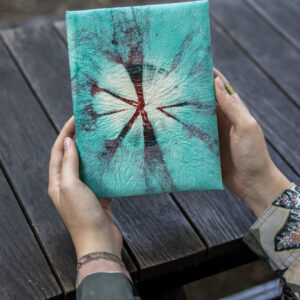 Osoba trzyma notes z turkusowo-bordowym wzorem tie-dye na drewnianym tle