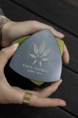Osoba otwiera duży czarno-złoty trójkątny grinder z logo Kana Sapiens na drewnianym tle