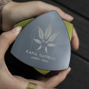 Osoba otwiera duży czarno-złoty trójkątny grinder z logo Kana Sapiens na drewnianym tle