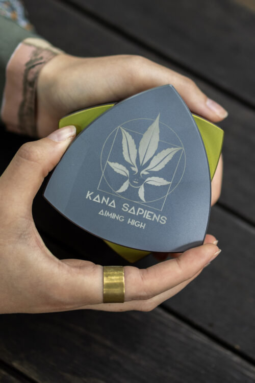 Grinder Osoba otwiera duży czarno-złoty trójkątny grinder z logo Kana Sapiens na drewnianym tle