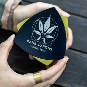 Grinder Osoba otwiera duży czarno-złoty trójkątny grinder z logo Kana Sapiens na drewnianym tle