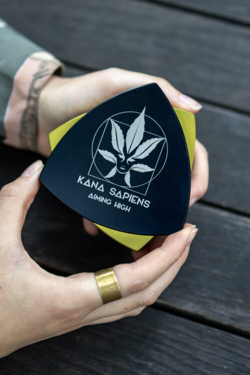 Grinder Osoba otwiera duży czarno-złoty trójkątny grinder z logo Kana Sapiens na drewnianym tle